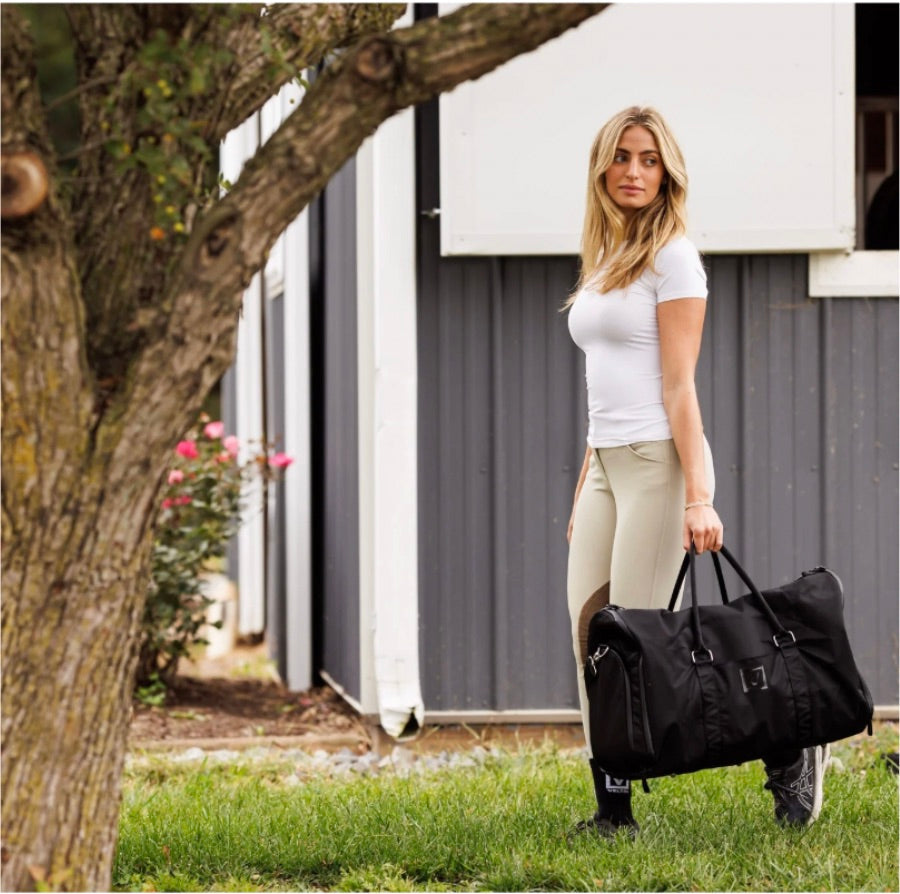 Woman in equestrian attire carrying a black Veltri Sport Ellis garment duffel bag outside a stable.