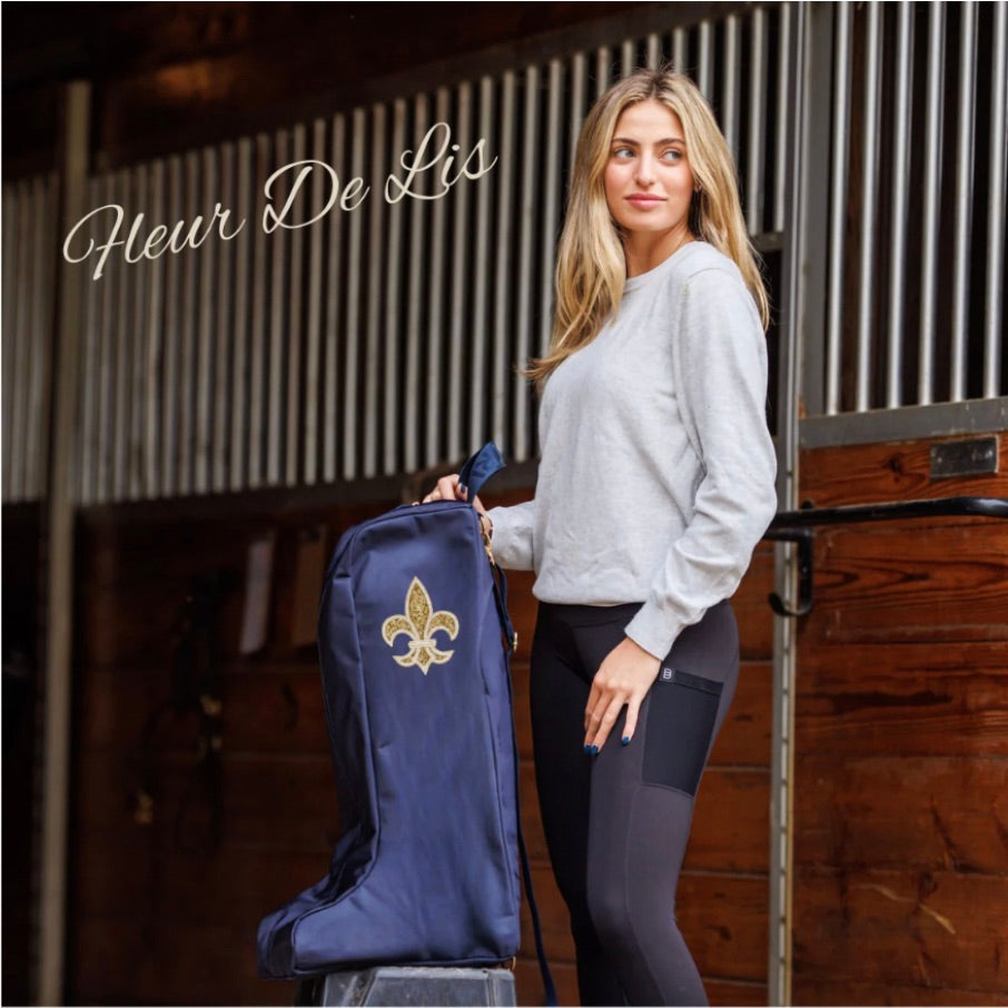 Woman in a stable holding a navy Veltri Sport Bedford boot bag with a gold fleur-de-lis embroidered, from the Fleur De Lis collection.