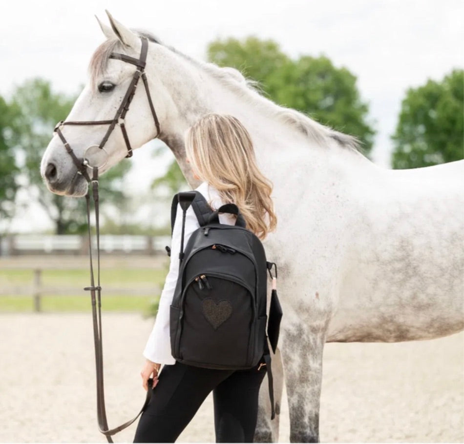 Woman wearing a black Veltri Sport Delaire backpack with a glitter heart embroidered standing beside a gray horse in an equestrian arena. 
