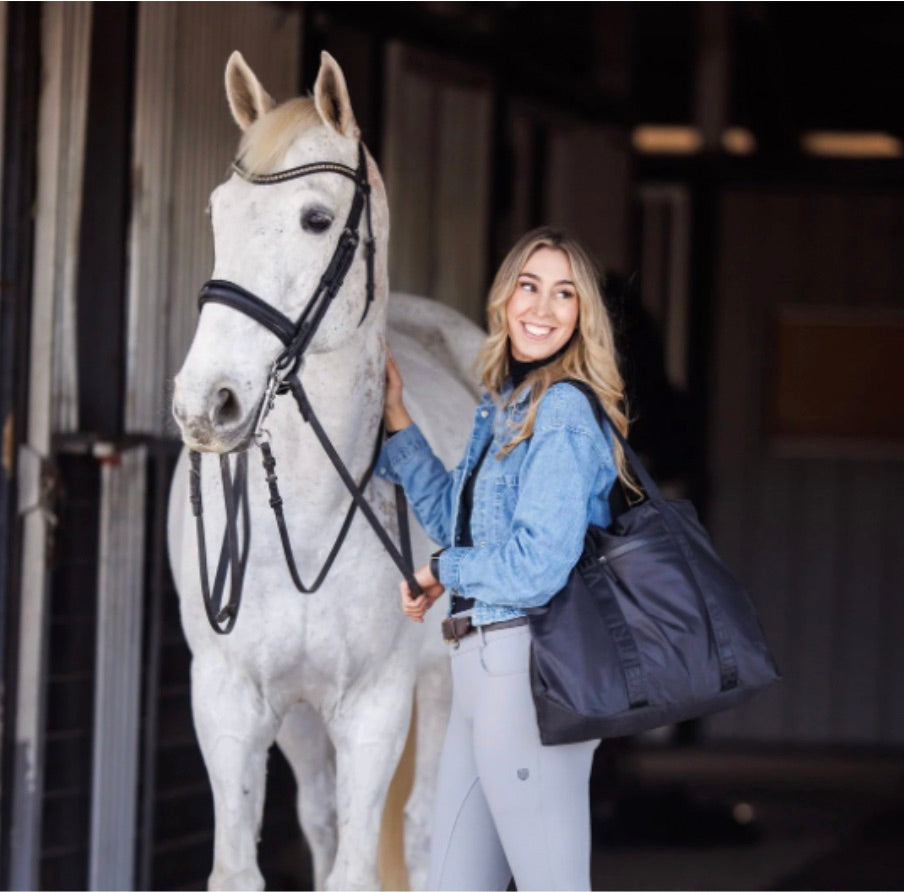 Smiling woman with a black Veltri Sport Newport tote bag standing beside a white horse at a stable. 
