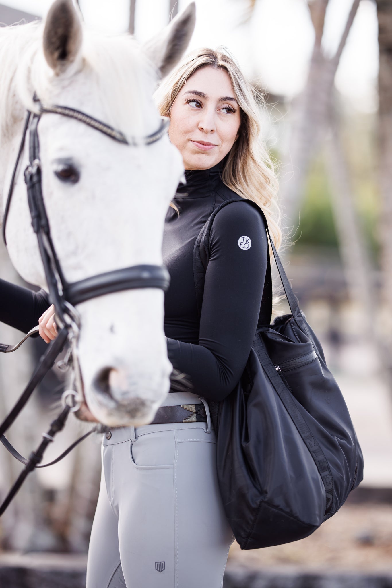 A horse mom is holding a white horse while carrying her Veltri Sport Newport Tote in black which has all of her and her daughter's horse show essentials.  