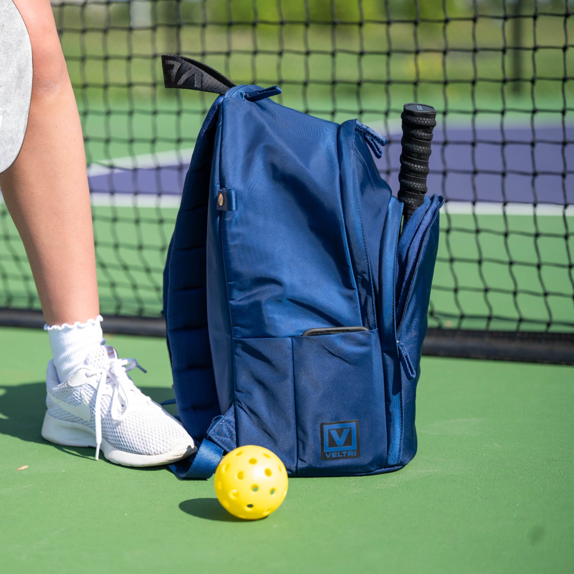 Veltri Sport Delaire Backpack standing courtside next to pickleball paddle and ball.