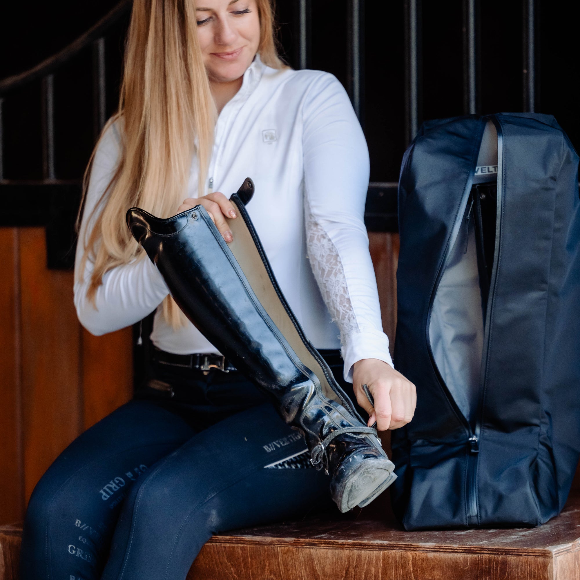 Woman sitting on a wooden bench putting equestrian boots in Veltri Sport Bedford Boot Bag.